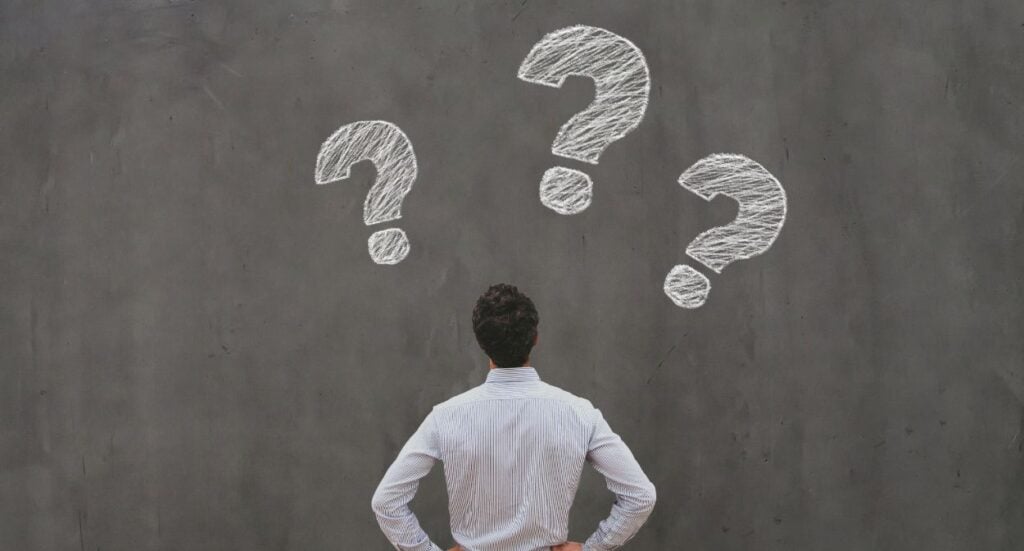 A person with brown hair in a white shirt stands facing a chalkboard with three large question marks drawn above their head, suggesting confusion or contemplation.