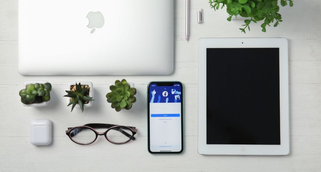 A flat lay of a workspace with a closed MacBook, an iPad, an iPhone showing a login screen, eyeglasses, AirPods, three small potted succulents, a pen, and a green plant on a white wooden surface.