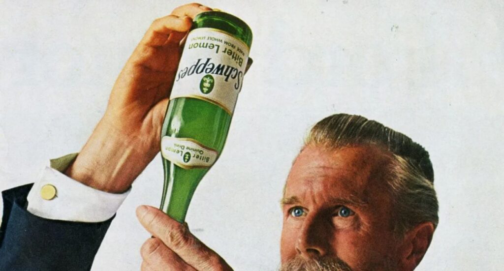 A man with a mustache and slicked-back hair holds a green Schweppes bottle upside-down, inspecting it closely against a plain light background.