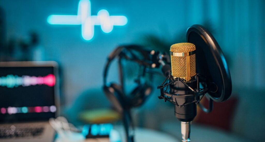 A gold microphone with a pop filter and headphones on a table, in front of a blurred laptop and neon waveform light in the background, creating a podcast or recording studio vibe.