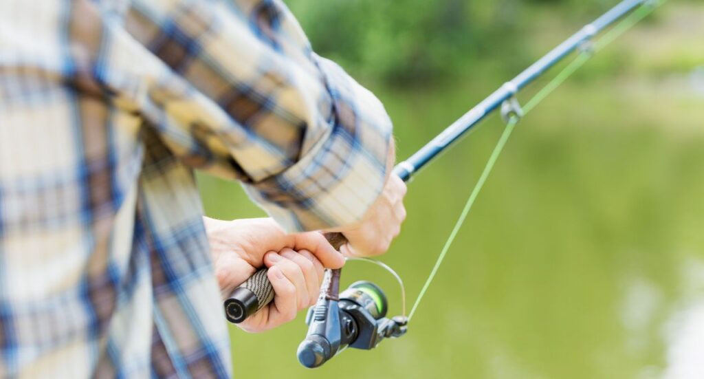 A person wearing a plaid shirt is holding and casting a fishing rod by a body of water, with a green, blurred background.