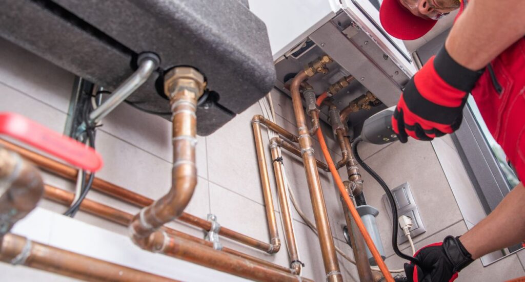 A plumber wearing red gloves and a cap repairs copper pipes connected to a wall-mounted boiler or water heater in a utility room. Various pipes and fittings are visible against the tiled wall.