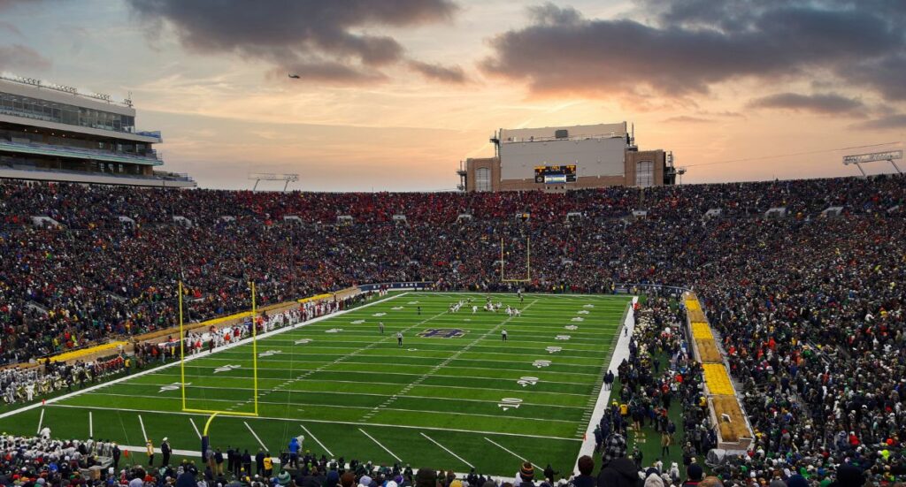 Packed football stadium at sunset with a game in progress, fans filling the stands, teams on the field, and a dramatic sky in the background.