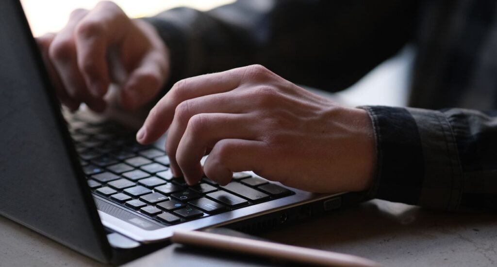 Close-up of a person’s hands typing on a laptop keyboard, with a pen lying on the table nearby. The person is wearing a dark plaid shirt.