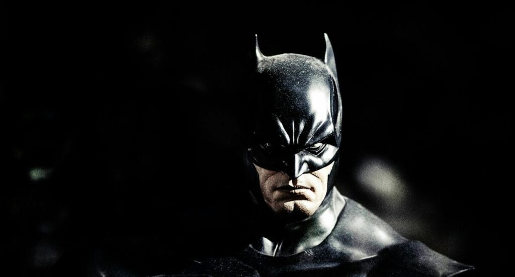 A close-up of Batman wearing his signature black cowl and cape, with a serious expression on his face, against a dark, shadowy background.