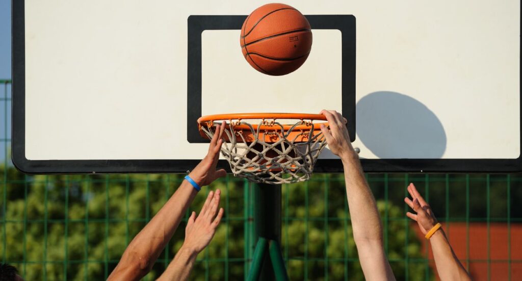 Three outstretched arms reach up toward a basketball approaching the hoop outdoors, with a green fence and trees blurred in the background.