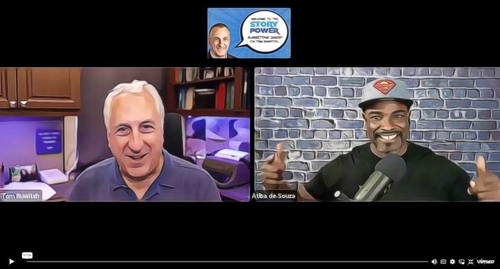 Two men are shown in a split-screen video call. The man on the left is smiling in an office, while the man on the right, wearing a Superman cap, smiles and gestures with both thumbs up in front of a brick wall and microphone.