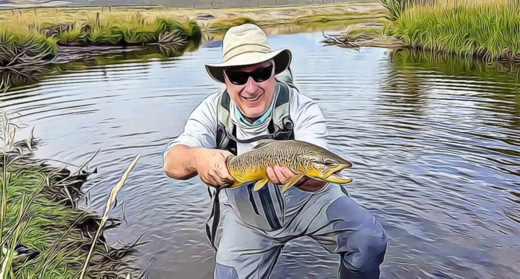 A person wearing a hat, sunglasses, and waders smiles while kneeling in shallow water, proudly holding a large brown fish, likely a trout, with grassy riverbanks and fields in the background.