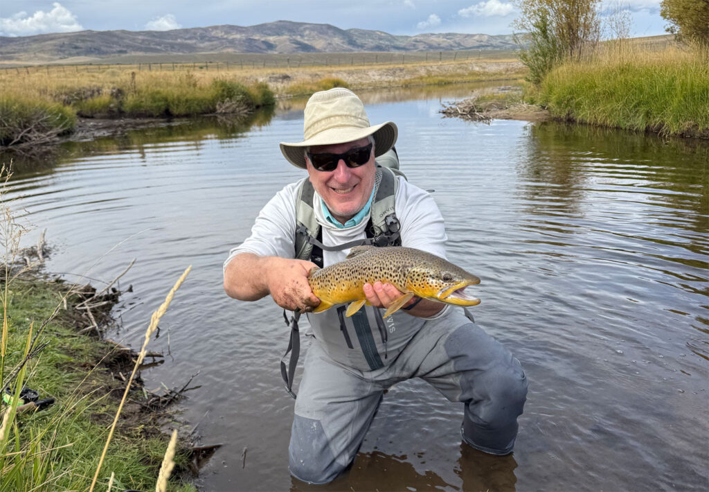 A smiling man in sunglasses and a hat kneels in a shallow stream, proudly holding a large brown trout, with grassy banks and hills in the background under a partly cloudy sky.