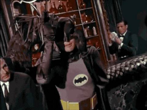 A person dressed as Batman in a classic costume performs a lively dance in a bar or club, while other people in suits watch and a bartender stands behind the counter.