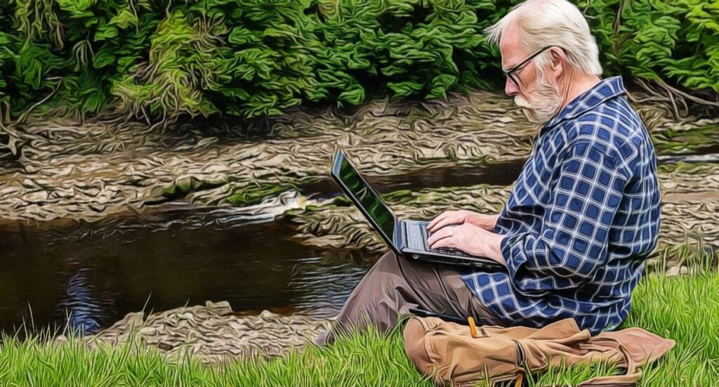 An older man with white hair and a beard sits on grass by a river, typing on a laptop. He wears a blue plaid shirt and glasses. A brown bag rests beside him, and green trees line the background.