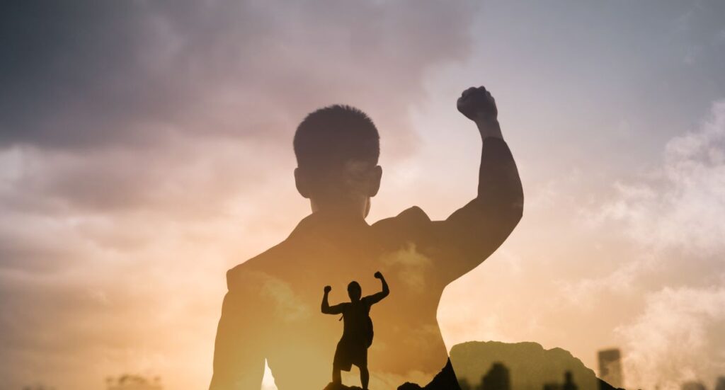 A silhouette of a person raising a fist in triumph is overlaid with a larger silhouette of the same pose, set against a cloudy sky at sunrise or sunset, symbolizing achievement and confidence.