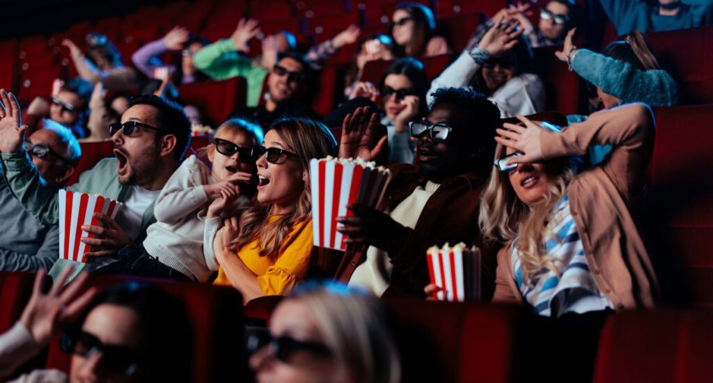 A group of people in a movie theater wearing 3D glasses react with surprise, holding popcorn and raising their hands as they watch the screen. The audience appears excited and engaged.