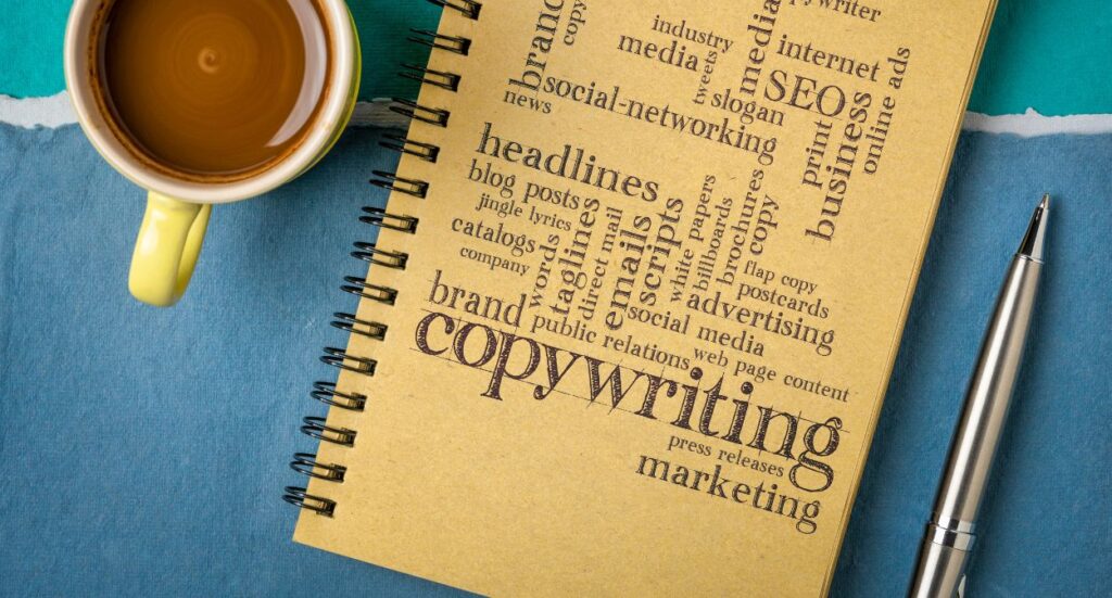 A yellow coffee cup, a silver pen, and a spiral notebook with the word copywriting and other marketing-related terms printed on the cover, all placed on a blue surface.