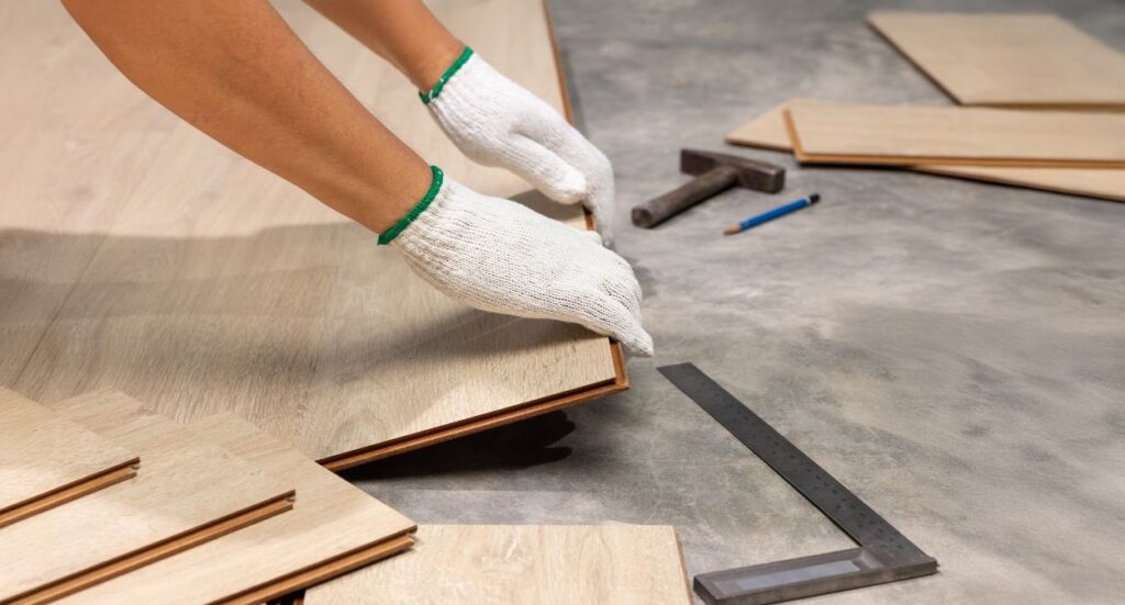 A person wearing white gloves installs wooden laminate flooring on a concrete surface, with tools like a hammer, ruler, and pencil scattered nearby.