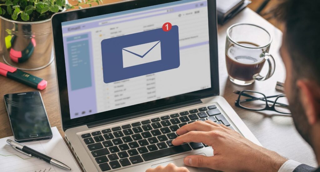 A person using a laptop sees a new email notification on the screen. There are office supplies, a plant, and a cup of coffee on the desk nearby.