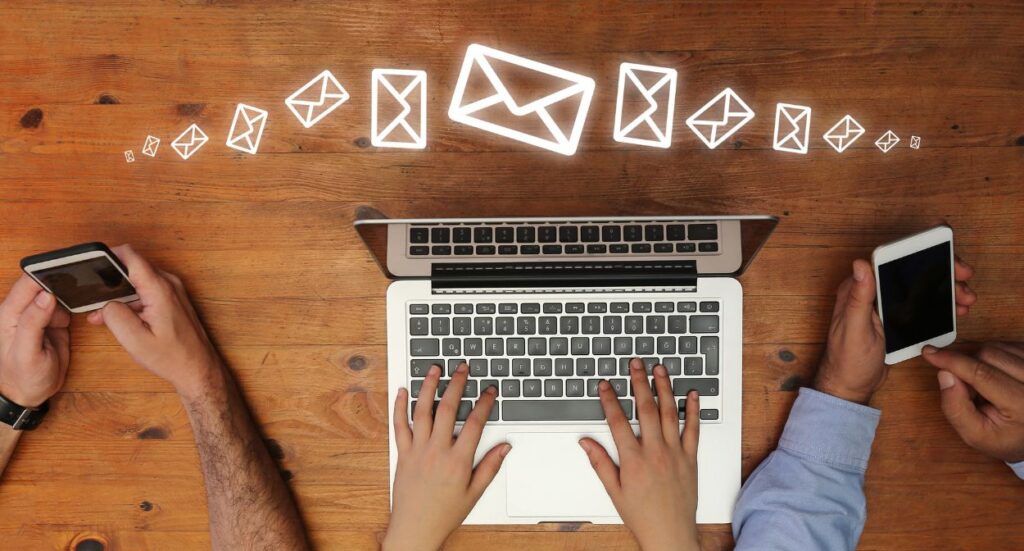 Three people use a laptop and smartphones on a wooden table, with digital email icons floating above the laptop, representing sending or receiving emails.