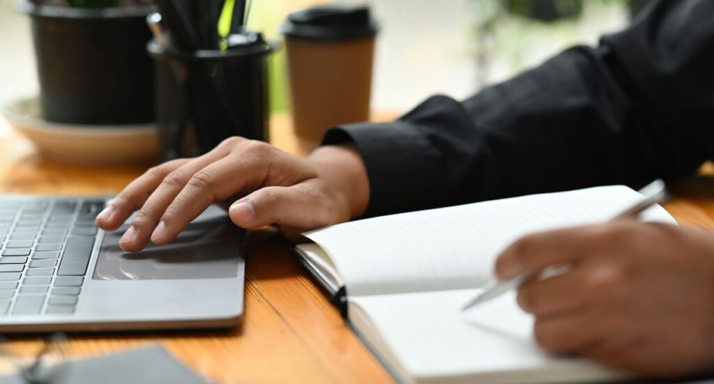 A person working at a desk with one hand on a laptop keyboard and the other writing in a notebook. A cup, pen holder, and other office items are visible in the background.