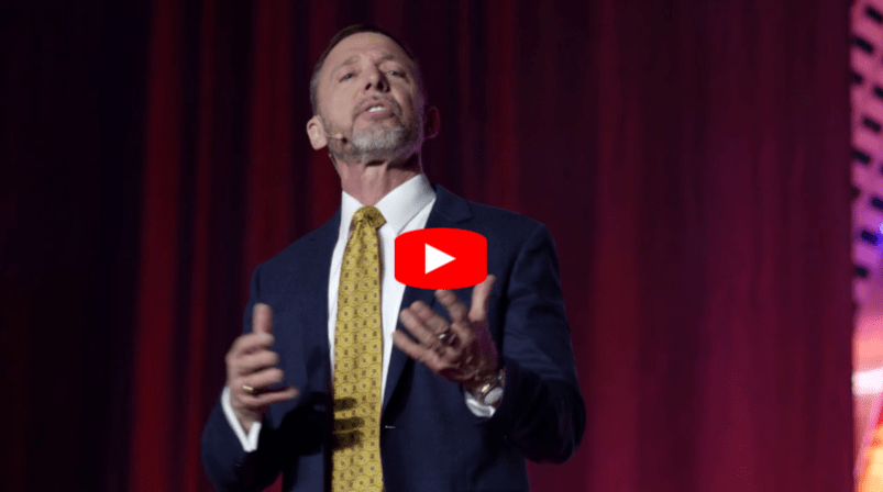A man in a dark suit and yellow patterned tie speaks on stage in front of red curtains, gesturing with his hands. A red YouTube play button icon is overlaid on the images center.