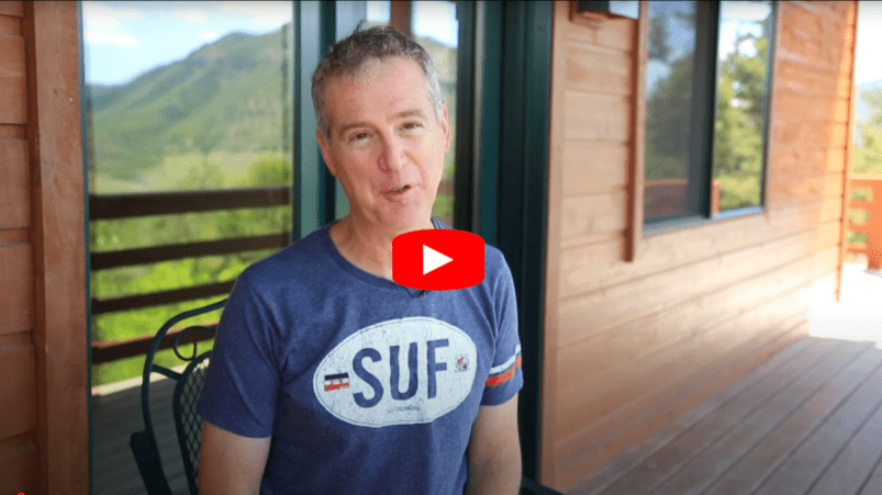 A man with short curly hair sits on a chair outside a wooden cabin, wearing a blue SUF T-shirt. Mountains and greenery are visible in the background. A red YouTube play button icon is overlaid on the image.