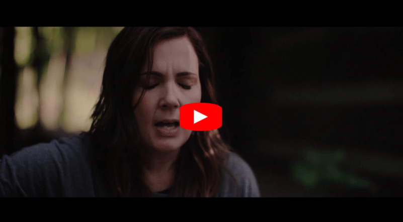 A woman with long brown hair sings with her eyes closed, wearing a gray shirt. The YouTube play button is overlaid on the image, suggesting it is a video thumbnail. The background is blurred with greenery visible.