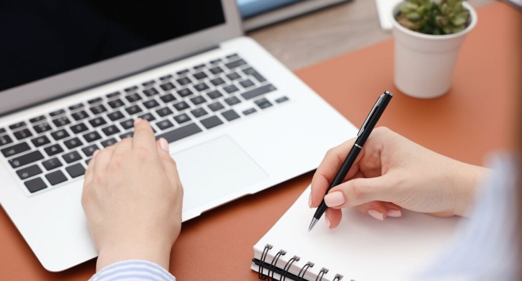 A person types on a laptop with one hand while holding a pen and writing in a spiral notebook with the other hand. A small potted plant is on the desk nearby.