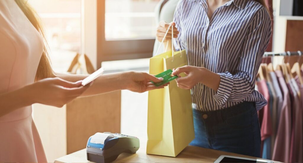 Two women are in a clothing store. One hands a green credit card to the other, who holds a yellow shopping bag. A payment terminal and tablet are on the counter in front of them. Sunlight comes through the window.