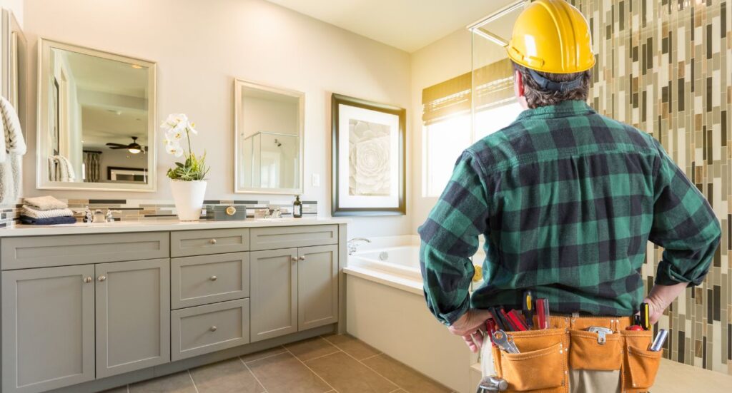 A construction worker in a yellow hard hat and tool belt stands in a modern bathroom, looking at the bathtub and vanity area with his hands on his hips.