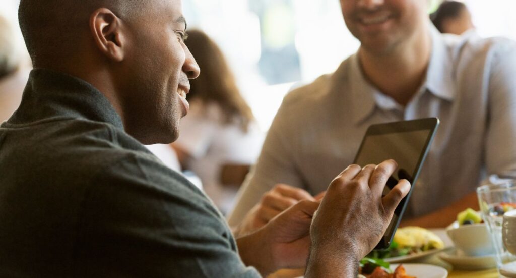 Two men sit at a table in a bright cafe, smiling and talking. One man holds and uses a tablet, while the other listens. Plates of food and drinks are visible on the table.