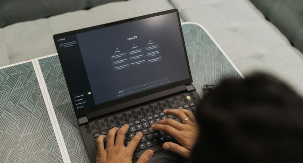 A person types on a laptop displaying the ChatGPT interface while sitting on a patterned surface with a gray cushion in the background.