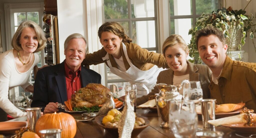 Five people smile around a dining table set for a meal, with a roast turkey as the centerpiece. The scene looks bright and festive, suggesting a holiday gathering, with pumpkins and autumn decorations visible.