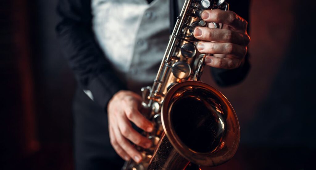 A person wearing a black suit and white shirt is playing a saxophone, with their hands on the instrument’s keys. The background is dark and out of focus.