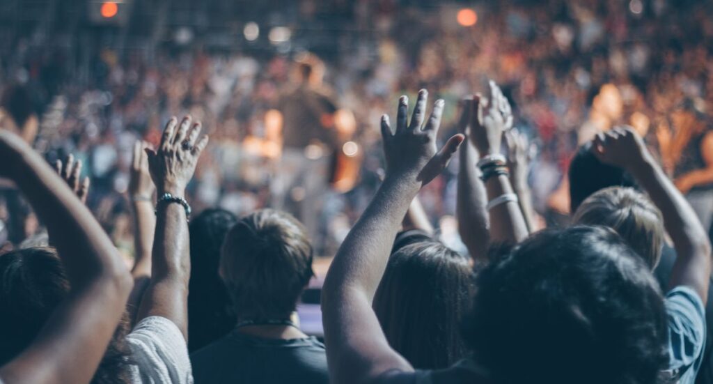A large crowd of people with raised hands faces a brightly lit stage, suggesting excitement and enthusiasm at a concert or live event. The scene is energetic and the audience is blurred in the background.
