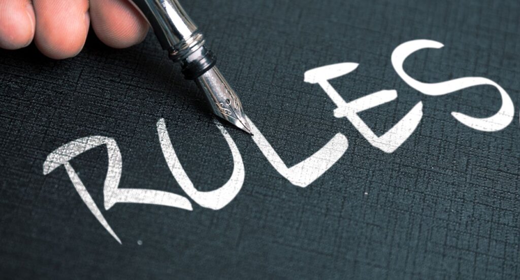 A close-up of a hand holding a fountain pen writing the word RULES in large, white, stylized letters on a textured black surface.