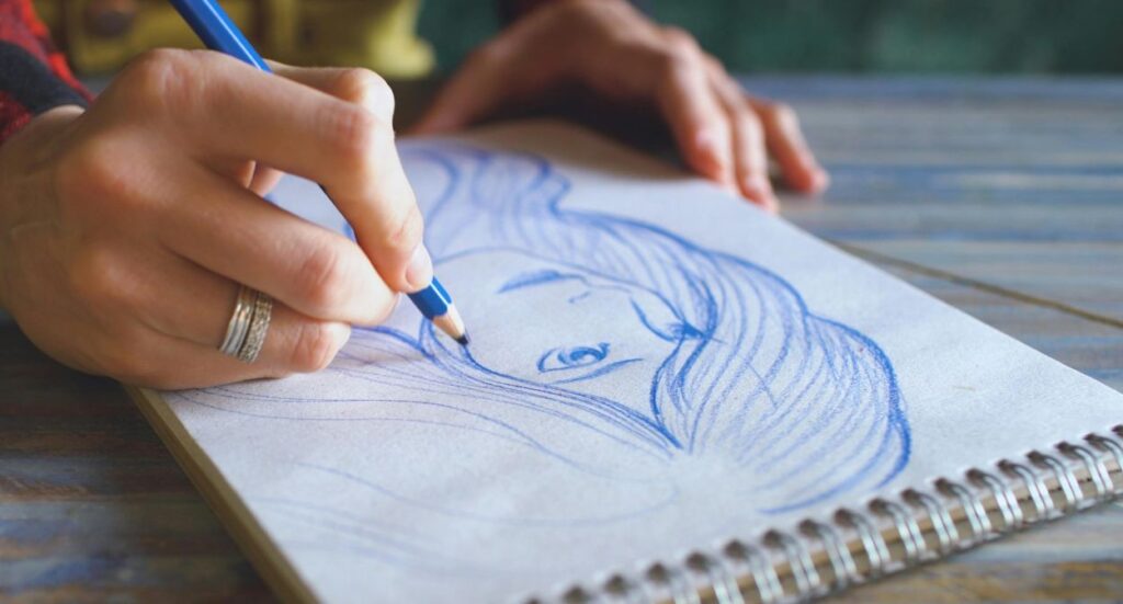 A person sketching a portrait of a woman with long hair in blue pencil on a spiral-bound sketchpad, their hand holding the pencil and another hand resting on the table.