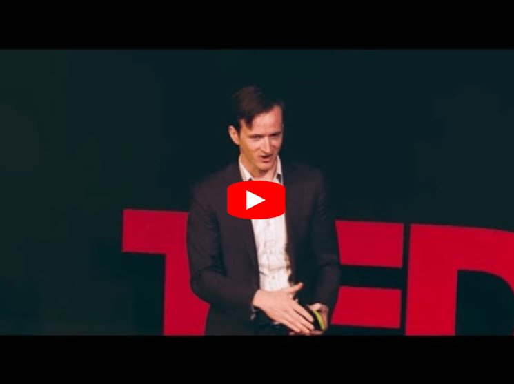 A man in a suit speaks on a stage with large red TED letters behind him, suggesting he is giving a TED Talk presentation.