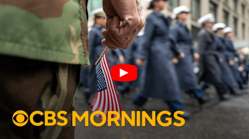 A person in camouflage holds a small American flag as uniformed individuals march past in the background; “CBS MORNINGS” is written at the bottom with a play button over the image.