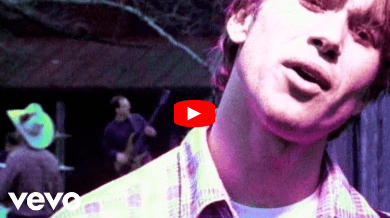 A close-up of a young man singing outdoors, with two musicians playing instruments in the blurry background. The lighting has a purple tint. The Vevo logo and a play button overlay are visible.