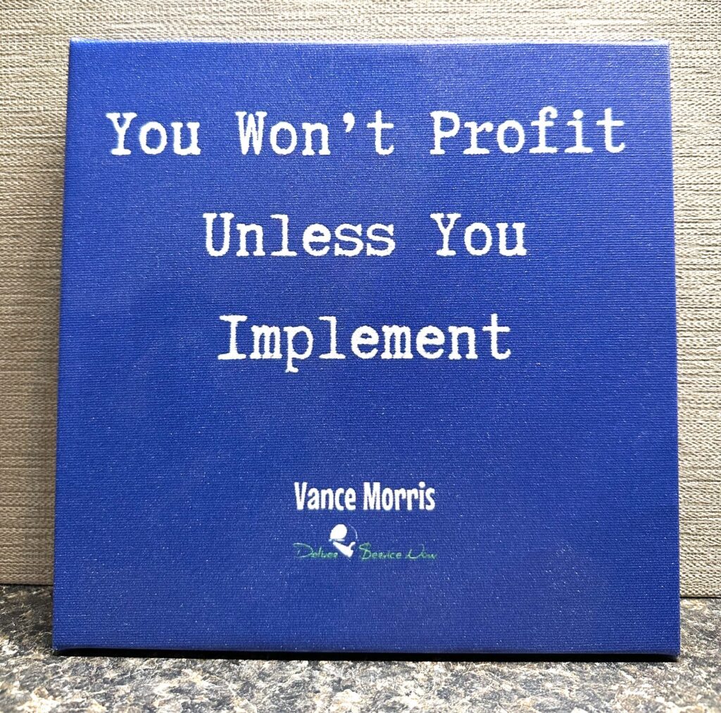 A blue sign with white text reads, You Wont Profit Unless You Implement. Below, it says Vance Morris and has a small logo with the words Deliver Service Now.