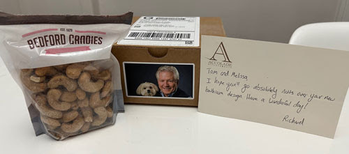 A bag of Bedford Candies cinnamon-glazed cashews, a box with a label featuring a man and a dog, and a handwritten note addressed to Tom and Melissa wishing them a wonderful day, all arranged on a white surface.