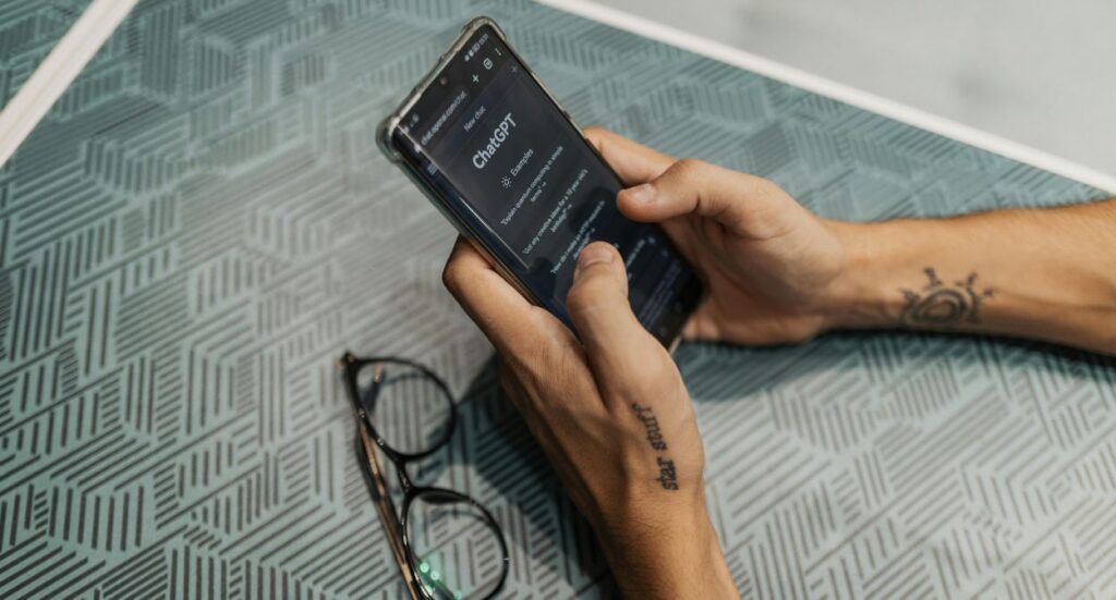 A person holding a smartphone with the ChatGPT website open, sitting at a patterned teal table. Their glasses and tattooed forearm are visible next to the phone.