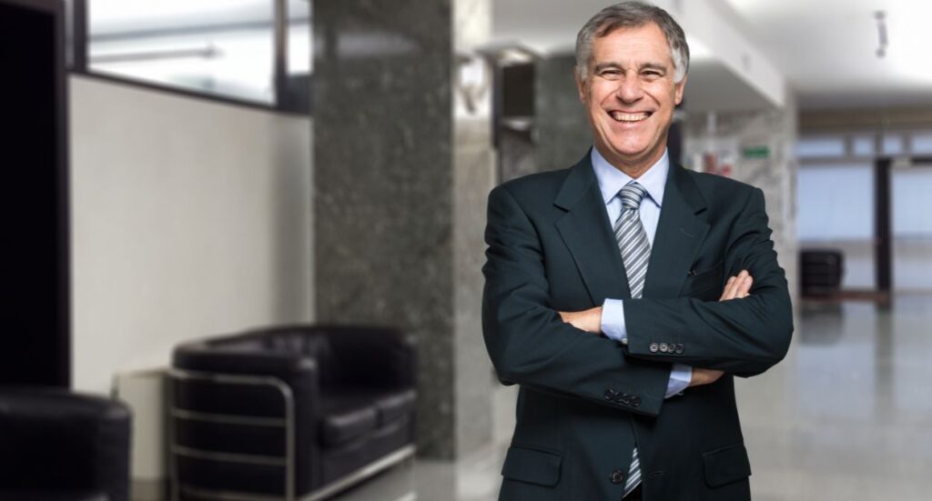 A smiling older man in a dark suit and striped tie stands with his arms crossed in a modern, bright office lobby with black chairs and marble walls in the background.