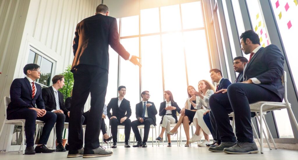 A group of professionals in business attire sit in a circle, listening to a standing presenter in a bright, modern office with large windows and sunlight streaming in.