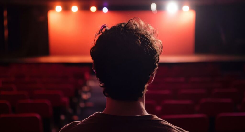 A person with short, curly hair is seen from behind, sitting alone in a theater with empty red seats and a brightly lit stage in the background.