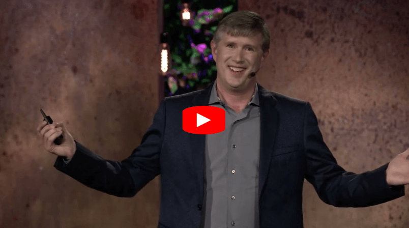 A man in a gray shirt and dark blazer stands on stage, smiling with arms open, holding a remote. Behind him is a textured brown wall with some colored lights. A large red YouTube play button is overlaid in the center.