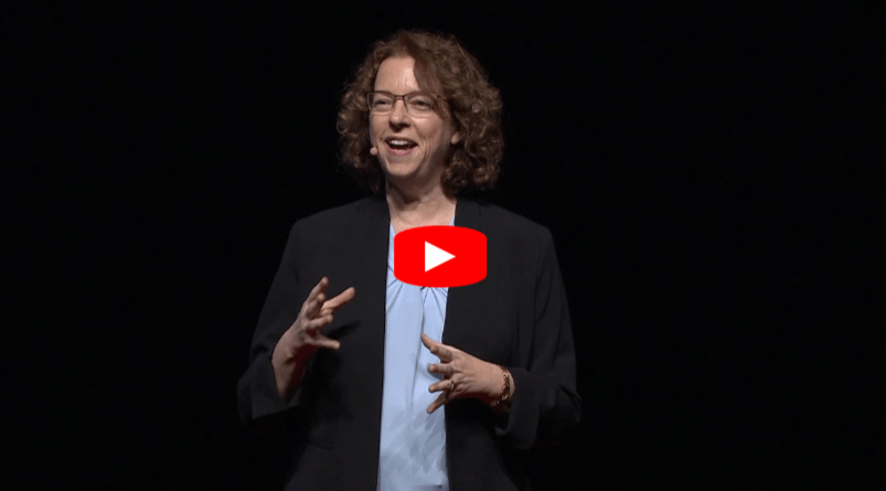 A woman with curly hair wearing glasses, a black blazer, and a light blue blouse speaks enthusiastically on stage against a dark background. A red YouTube play button is overlaid at the center of the image.