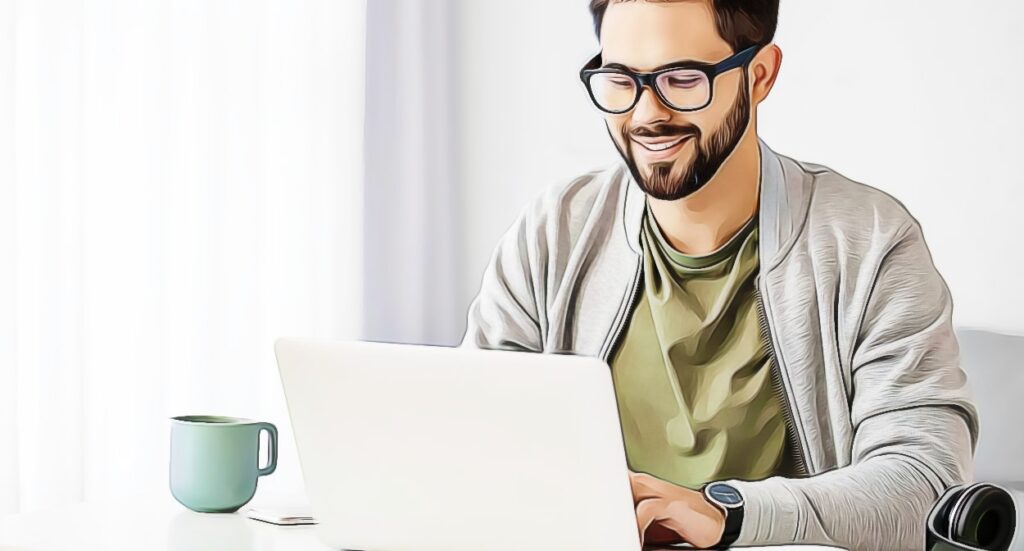 A smiling man with glasses and a beard works on a laptop at a desk. He wears a light jacket over a green shirt, with a coffee mug and headphones nearby in a brightly lit room.