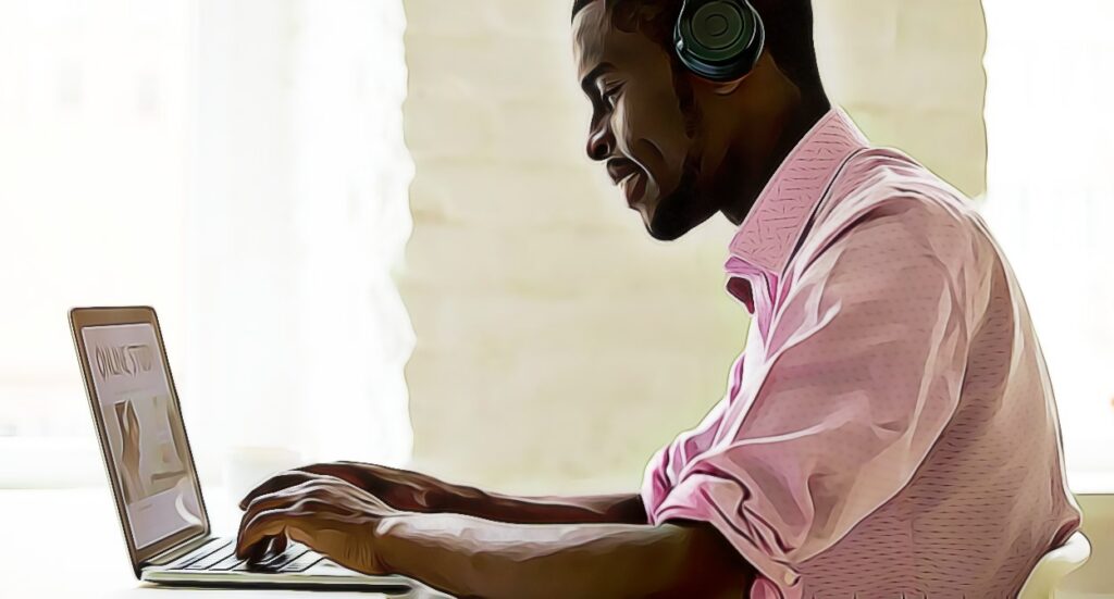 A man wearing headphones and a pink shirt sits at a desk, smiling while typing on a laptop in a brightly lit room.