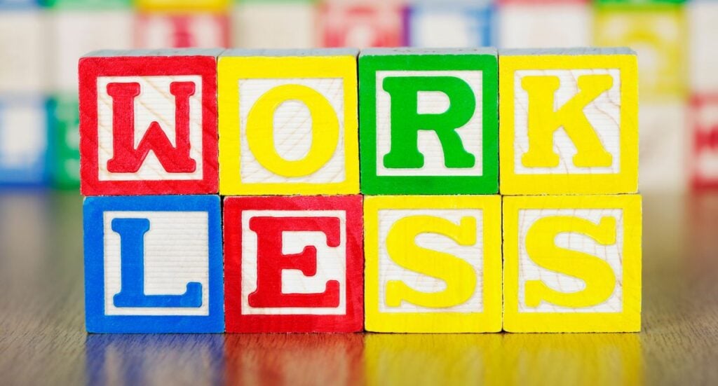 Colorful toy blocks arranged to spell the words WORK LESS on a wooden surface, with a blurred background of more blocks.