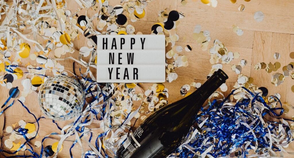 A “Happy New Year” sign sits on a wooden floor surrounded by a champagne bottle, a disco ball, confetti, and streamers in gold, silver, blue, and white.